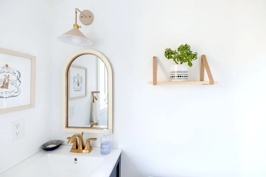 A small bathroom with a round mirror, wall shelf, and potted plant in warm natural light