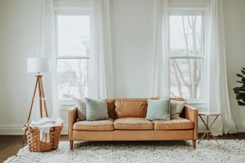 A warm minimalist living room with a cognac leather sofa, wood tripod floor lamp, and woven basket