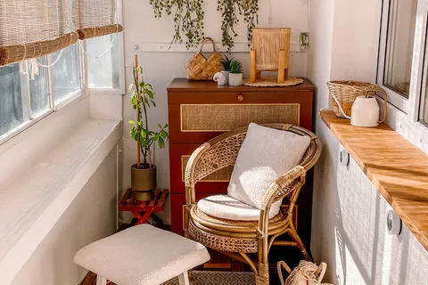 A small furnished apartment balcony with a rattan chair, hanging plants, and a patterned rug on a wood tile floor