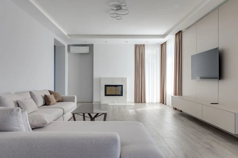 A modern minimalist living room with a gray sectional sofa, marble fireplace, and wall-mounted TV in warm neutral tones