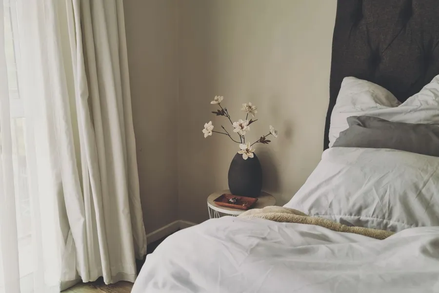 A calm, neutral bedroom with a curtained window and a black ceramic vase of white flowers on the nightstand