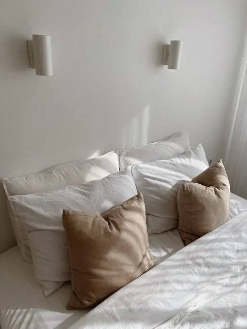A warm neutral bedroom with beige and white pillows against a soft white wall, an example of the best color for bedroom walls with little direct light