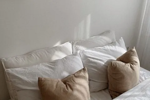 A warm neutral bedroom with beige and white pillows against a soft white wall, an example of the best color for bedroom walls with little direct light