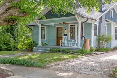A small blue craftsman house with a painted gray front porch, two white rocking chairs, and a wood front door seen from the street