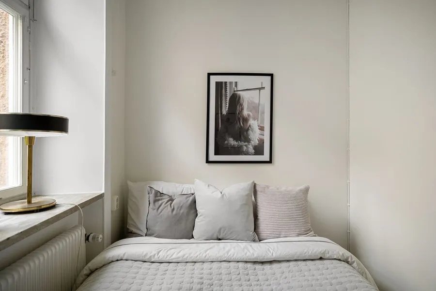A small, warm master bedroom with soft daylight, a table lamp, and layered neutral bedding