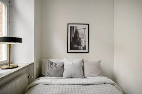 A small, warm master bedroom with soft daylight, a table lamp, and layered neutral bedding
