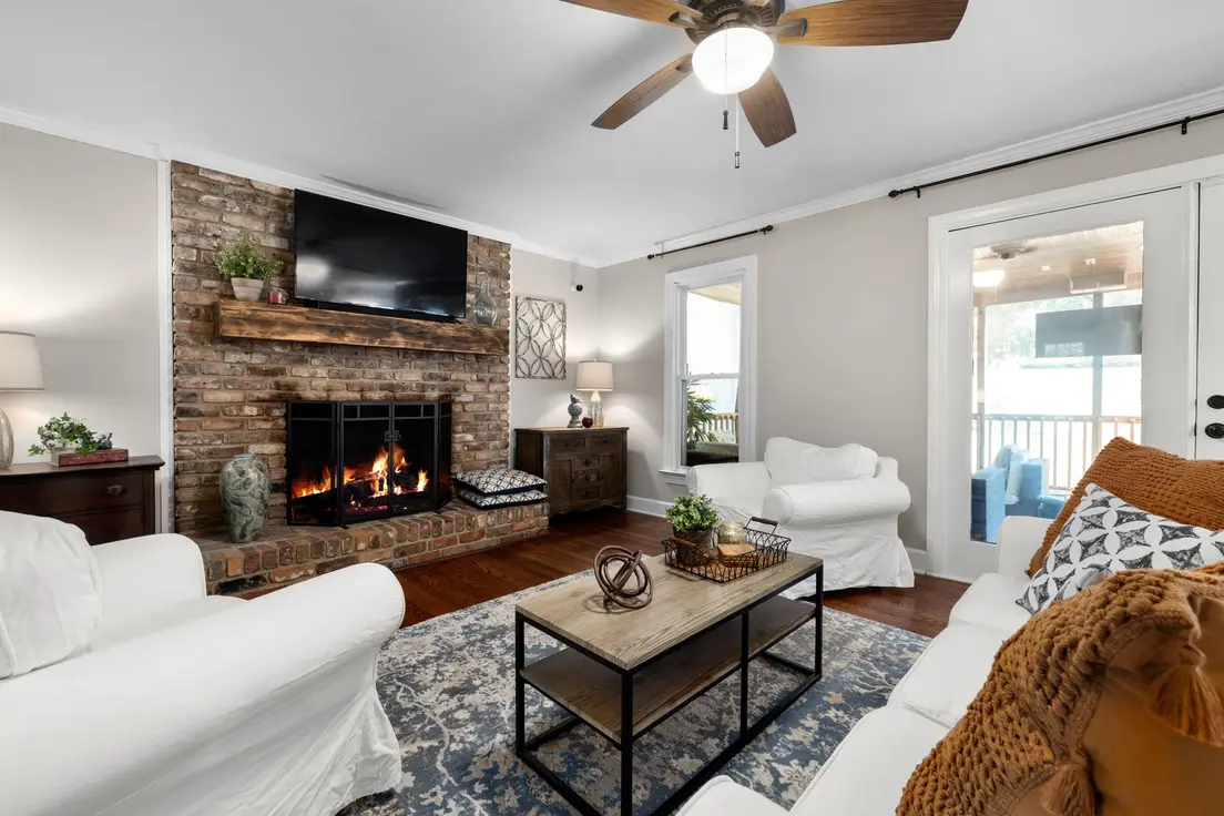 A modern farmhouse living room with white slipcovered armchairs, a brick fireplace with a rustic wood mantel, and a wood-and-black-metal coffee table