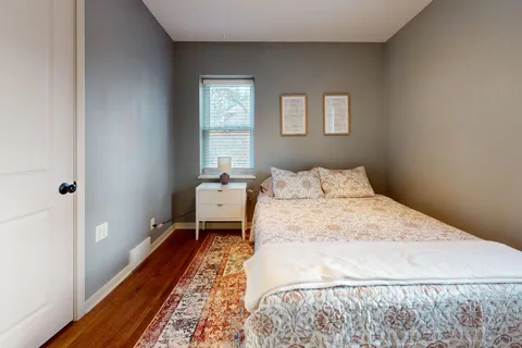 A small, pale gray-blue master bedroom with a patterned rug, white nightstand, and one window