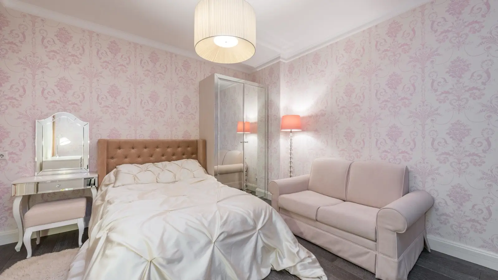 A feminine bedroom with pink floral wallpaper, a tufted brown headboard, a mirrored vanity, and a pendant light