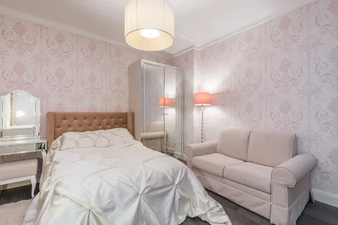 A feminine bedroom with pink floral wallpaper, a tufted brown headboard, a mirrored vanity, and a pendant light