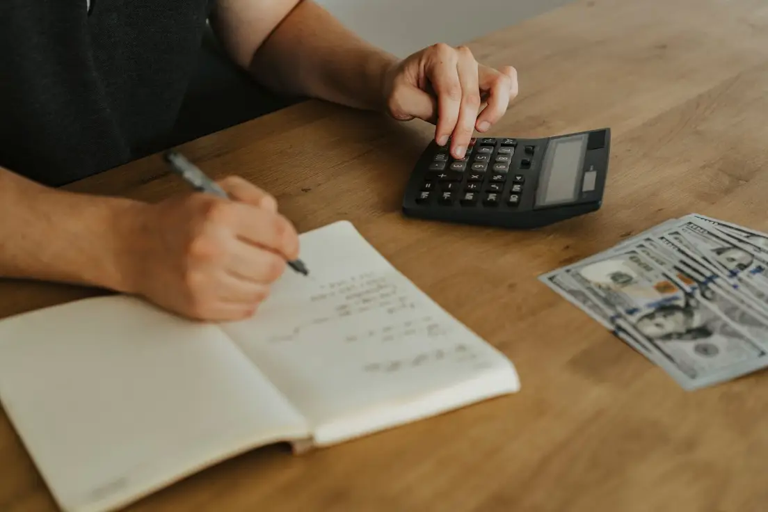 Hands working out a bedroom decor budget with a calculator, notebook, and cash on a wood table