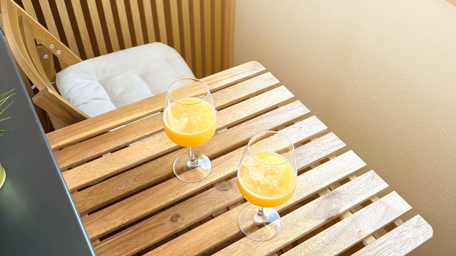 A small wooden balcony table with cushioned chairs and two glasses of juice in morning light