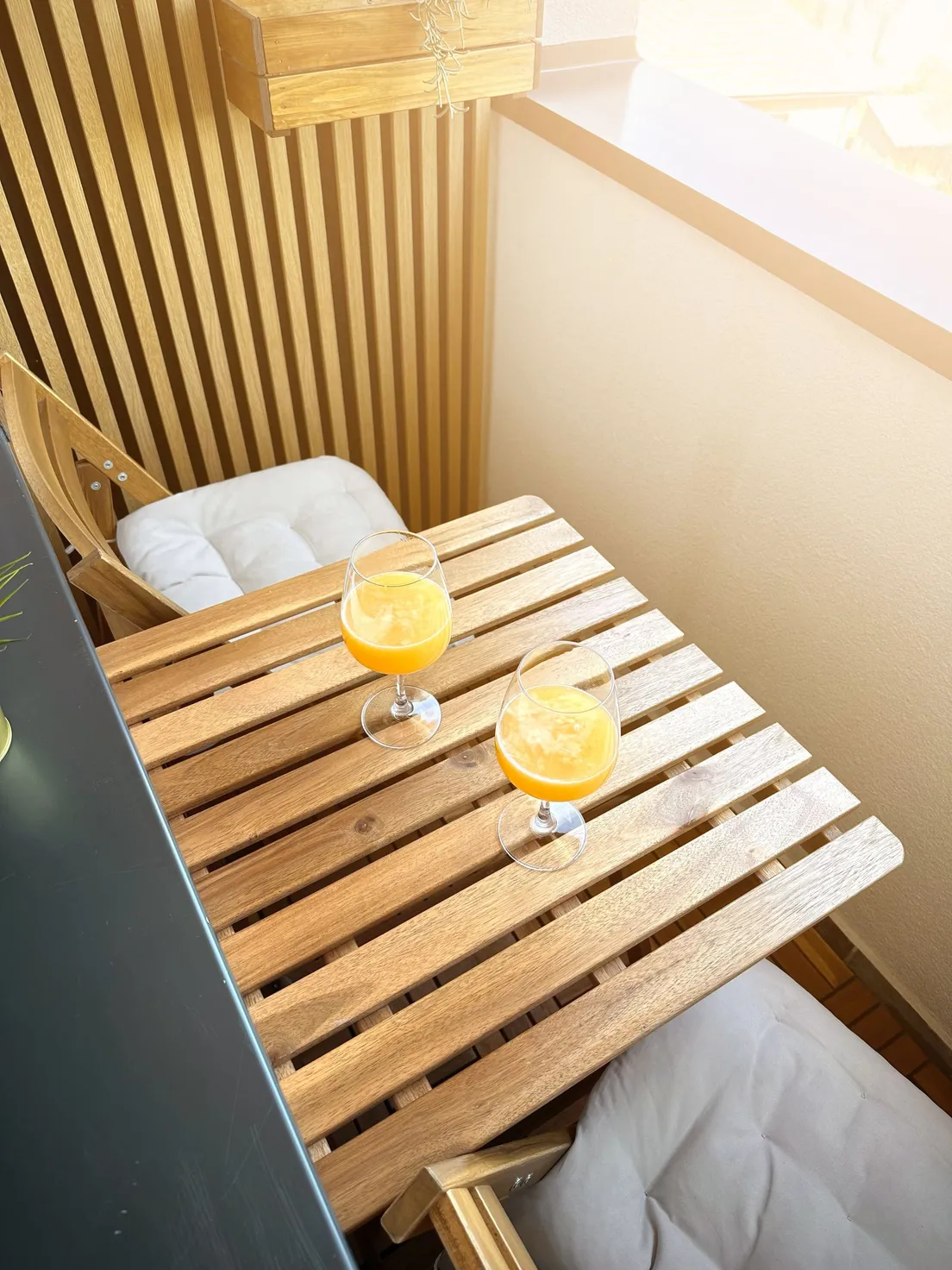 A small wooden balcony table with cushioned chairs and two glasses of juice in morning light
