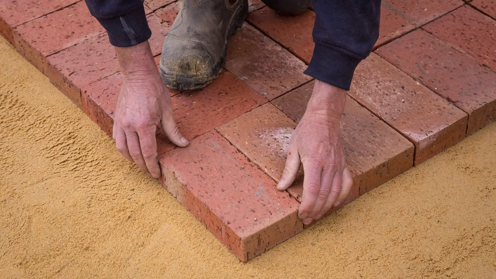 A worker's hands setting brick pavers onto a sand bed during patio installation
