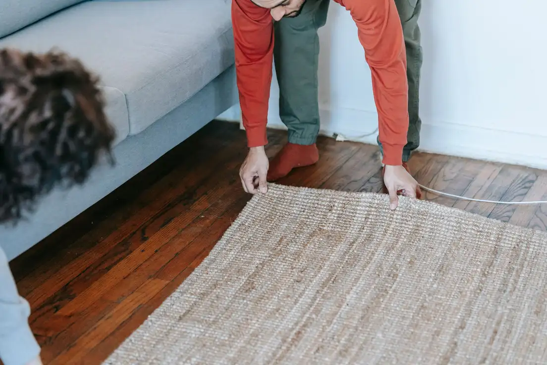 A person unrolling a natural jute rug across a dark wood floor beside a sofa in a farmhouse-style living room
