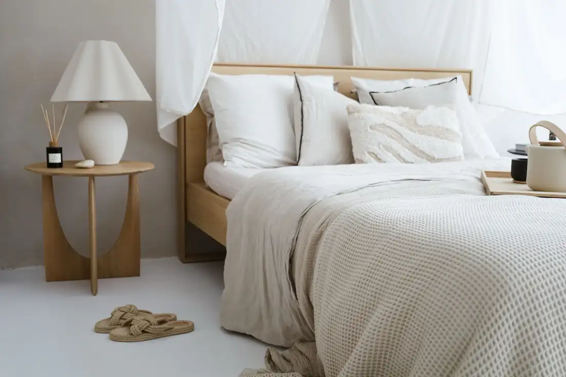 A bed made with layered neutral linen bedding, a waffle-knit throw, and a white ceramic lamp on a wood side table