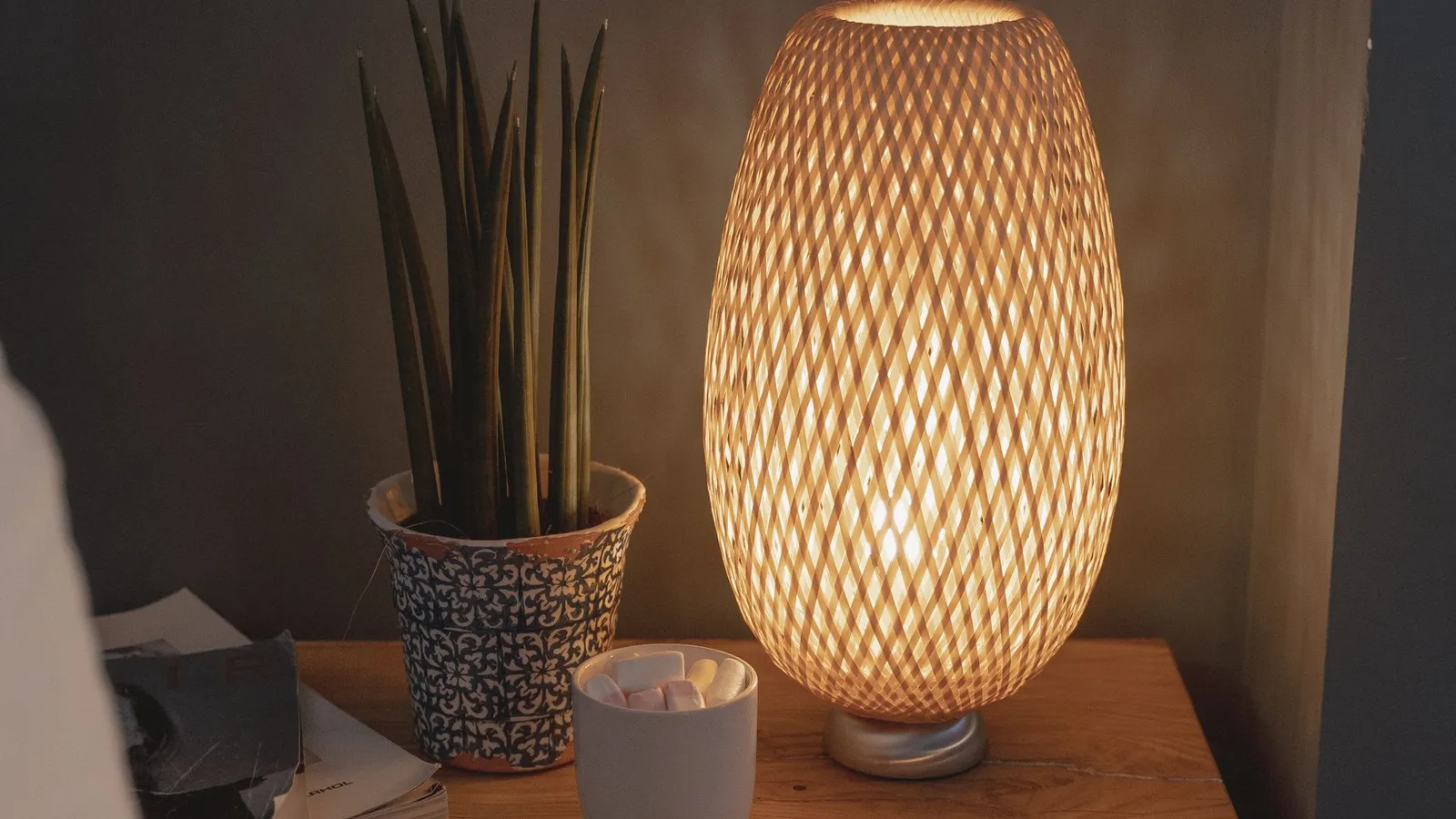 A woven bamboo table lamp glowing warmly on a wooden nightstand beside a small plant