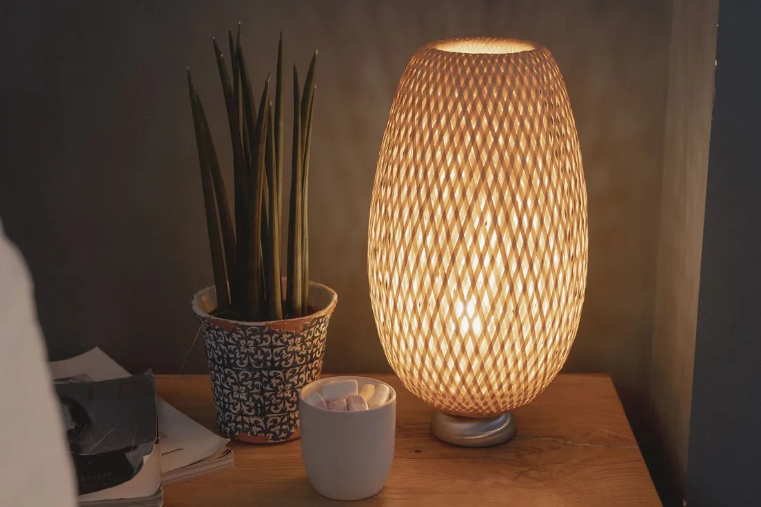 A woven bamboo table lamp glowing warmly on a wooden nightstand beside a small plant