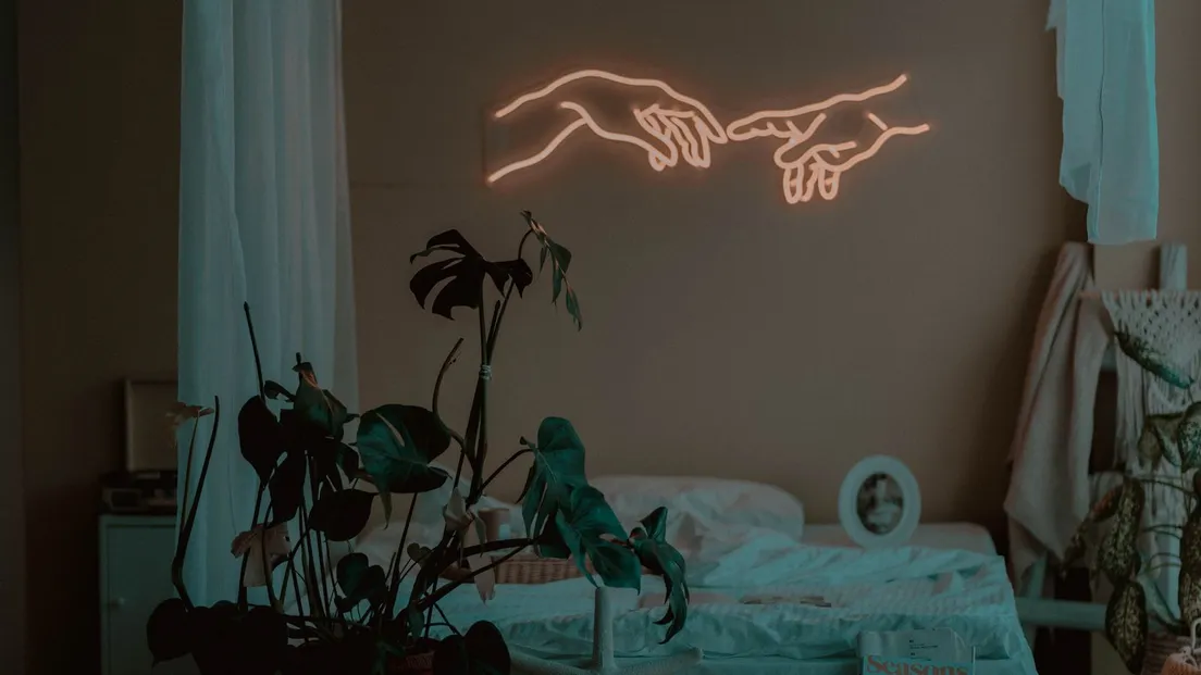 A dim bedroom lit by a warm coral neon sign shaped like hands above the bed