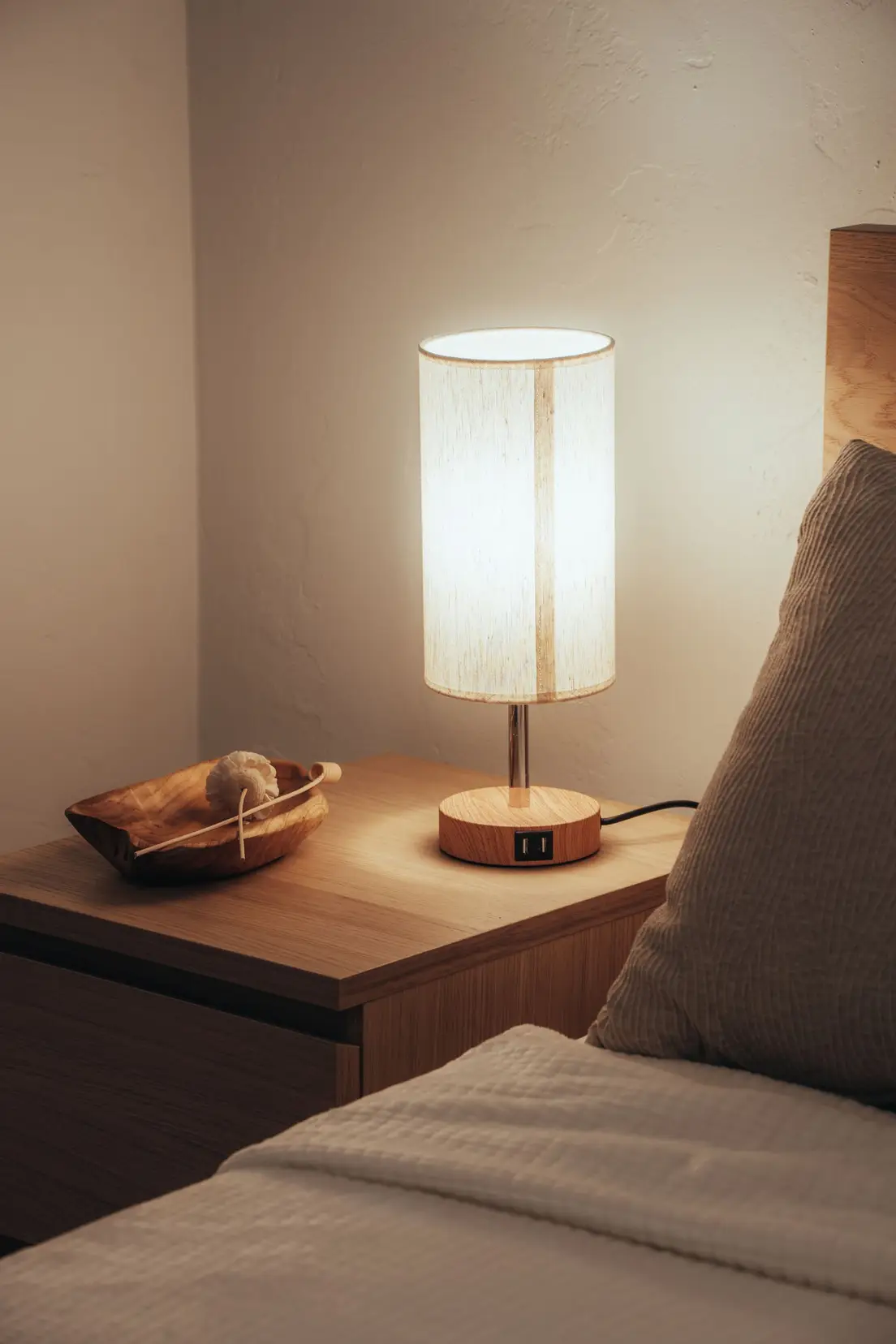 A warm wood table lamp glowing on a bedroom nightstand at night
