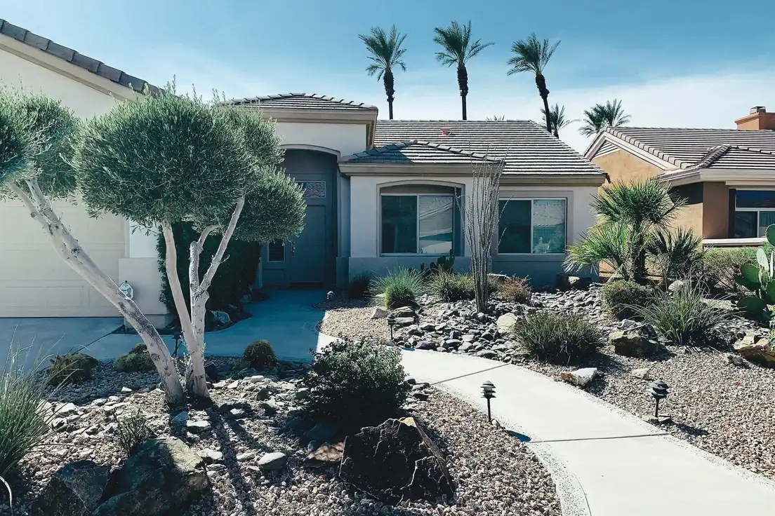 A low-maintenance desert landscape in front of a single-story ranch house with gravel, river rock, ornamental grasses, and a curved walkway