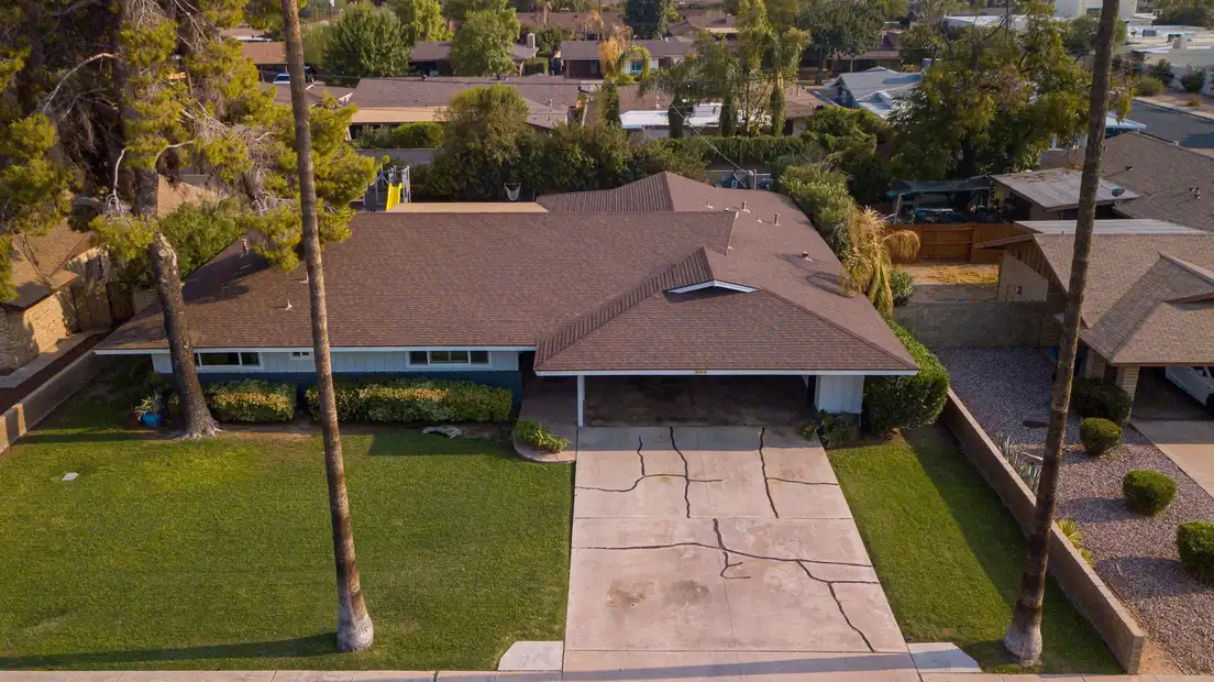 A mid-century ranch house seen from above, showing its low hip roof, deep eaves, and open carport over the driveway