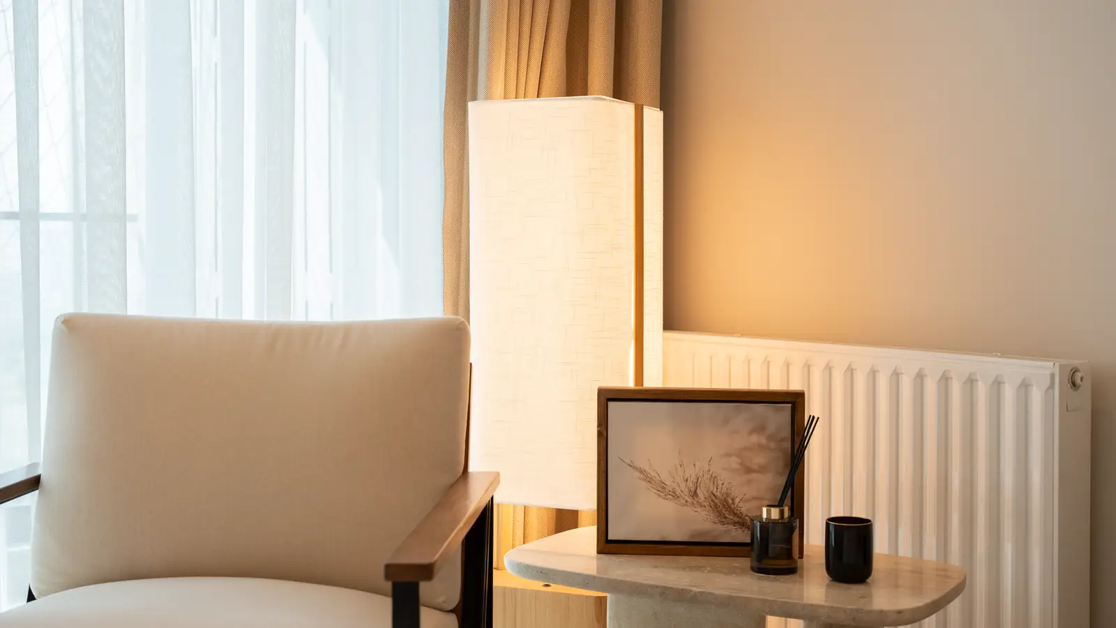 A warm minimalist reading nook with a cream armchair, linen lamp, and framed art on a marble side table
