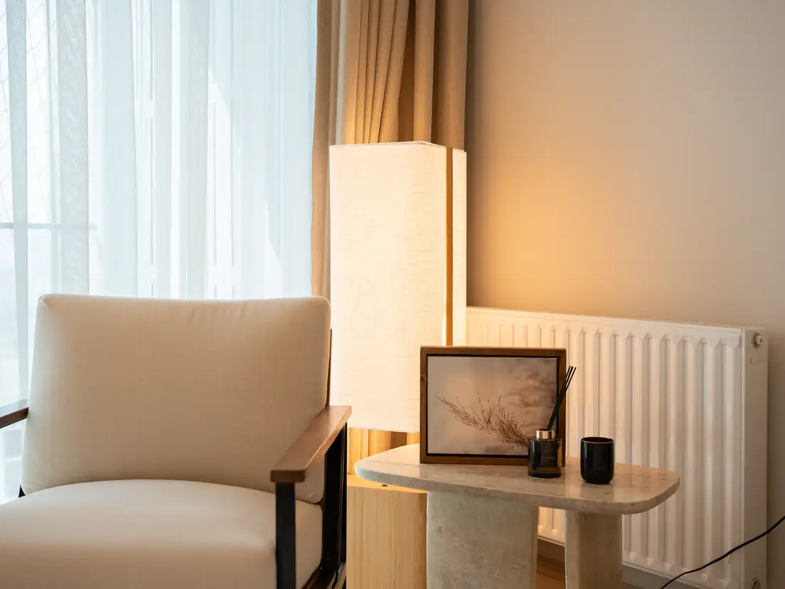 A warm minimalist reading nook with a cream armchair, linen lamp, and framed art on a marble side table