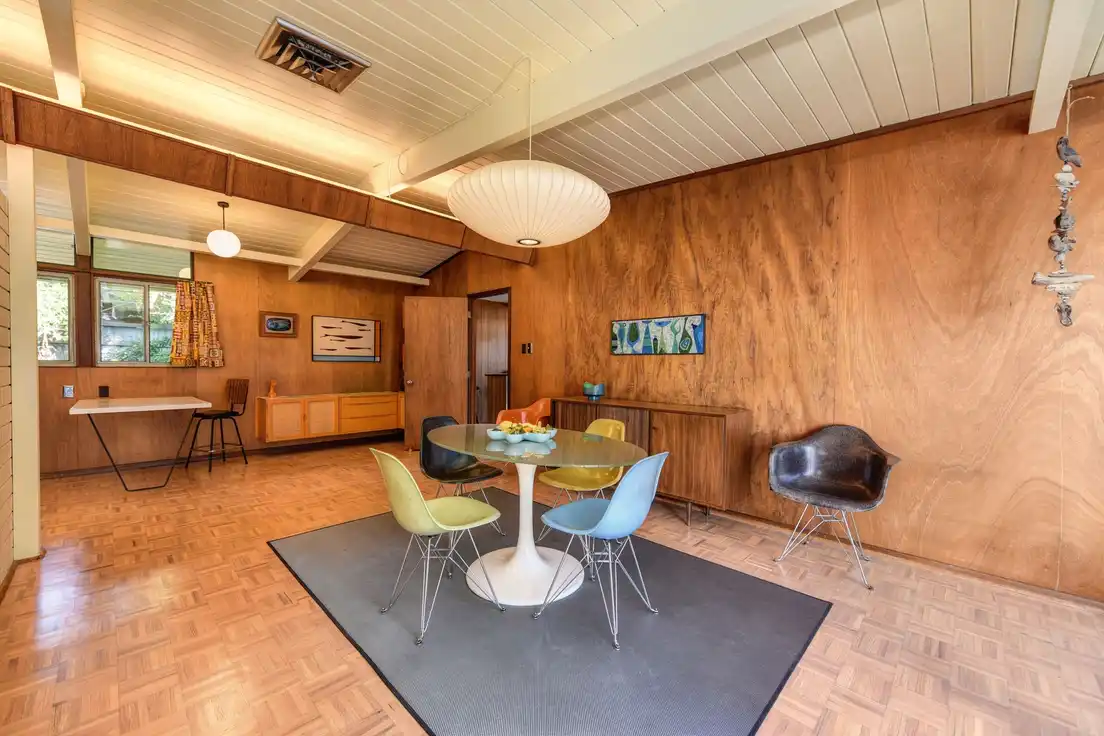 A mid-century dining room with wood-paneled walls, a round pedestal table, molded shell chairs, and a paper pendant lamp