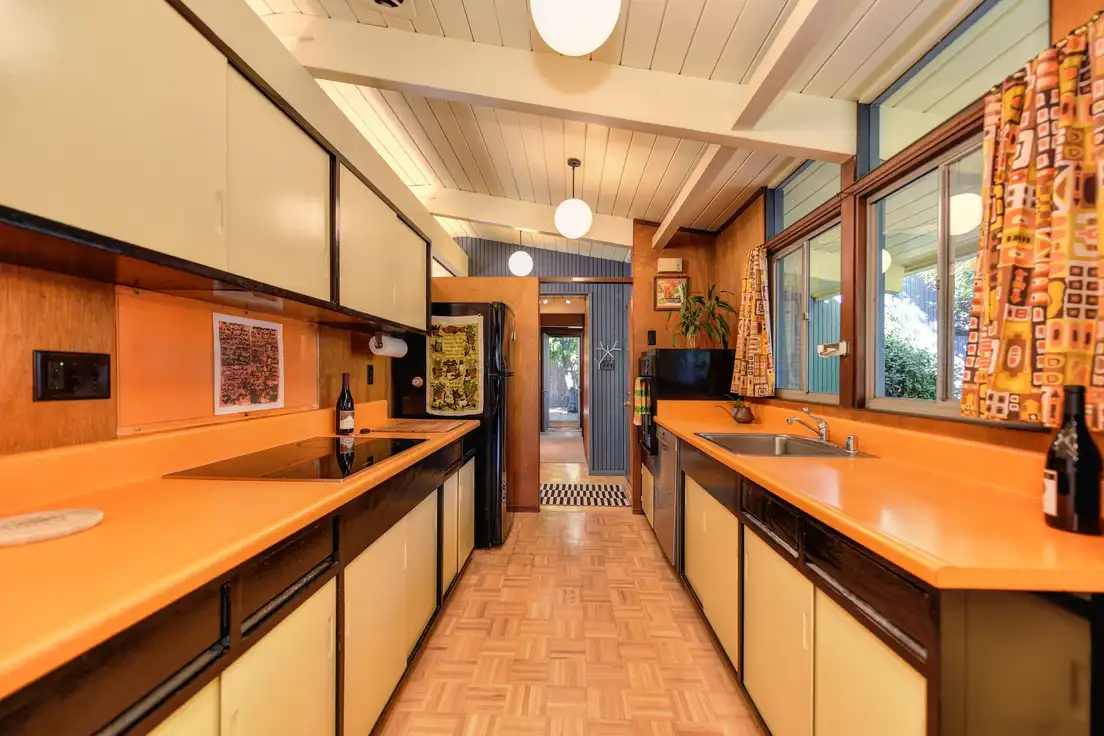 An original mid-century ranch galley kitchen with orange Formica countertops, cream sliding cabinet doors, a beamed ceiling, and globe pendants