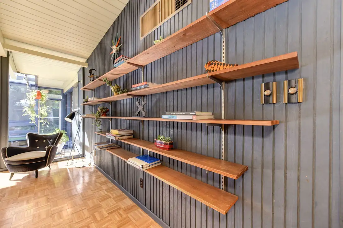 Inside a mid century modern ranch house: a gray board wall with long wood shelves, a parquet floor, and a glass wall to the yard