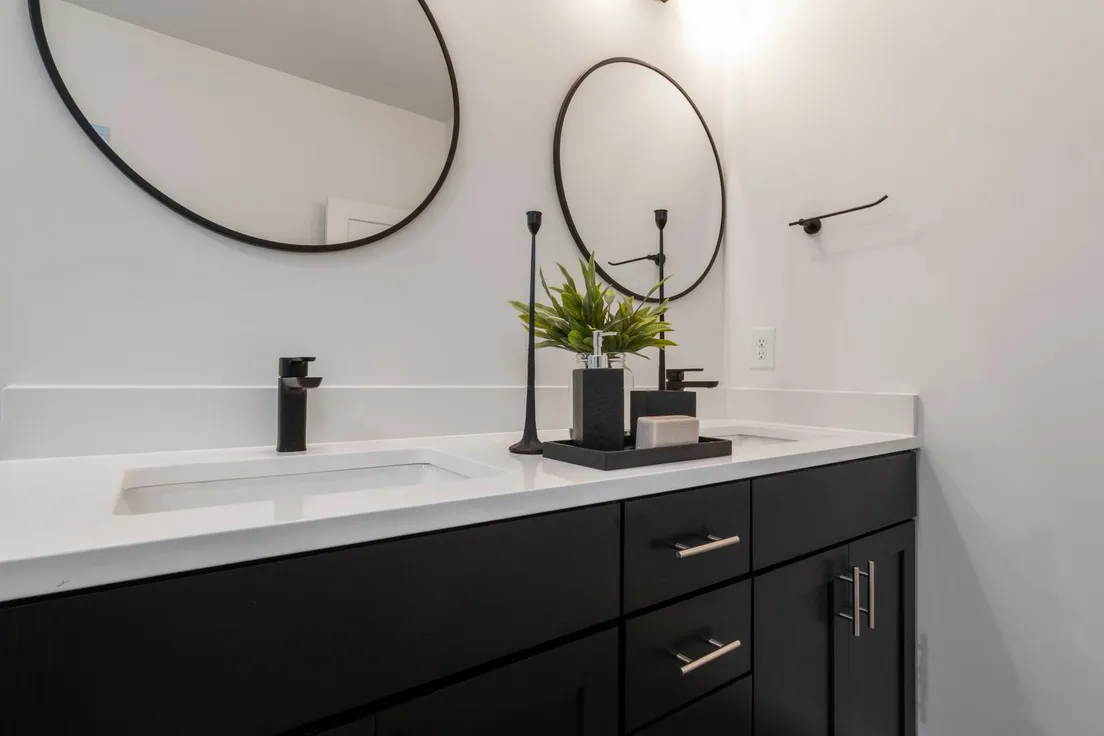Two round black-framed mirrors mounted above a dark bathroom vanity with a potted plant and candles between the sinks