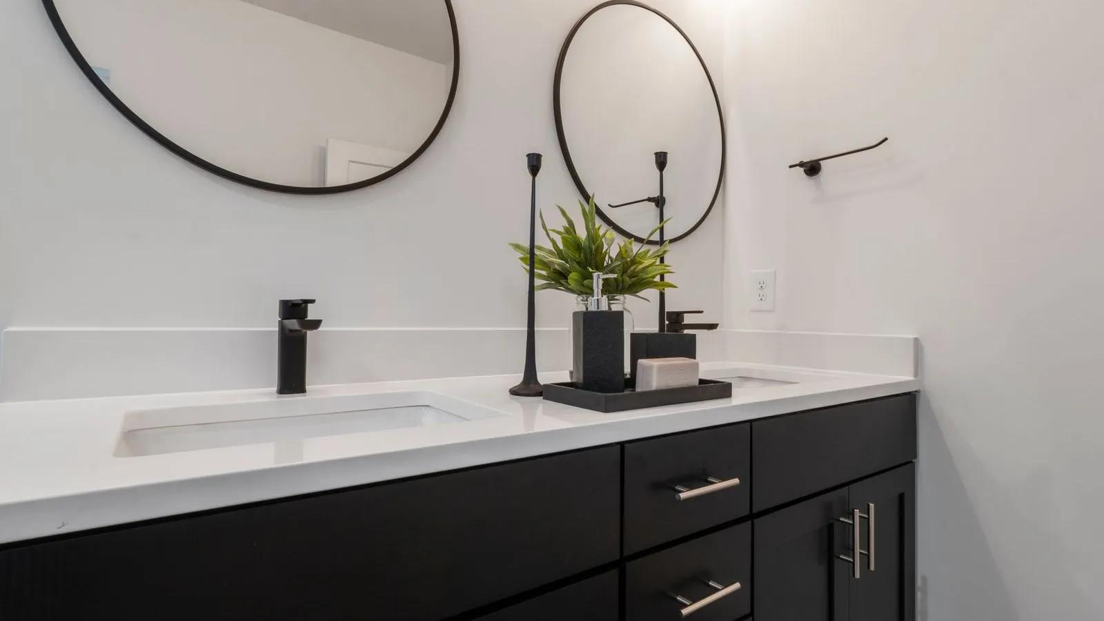 Two round black-framed mirrors mounted above a dark bathroom vanity with a potted plant and candles between the sinks