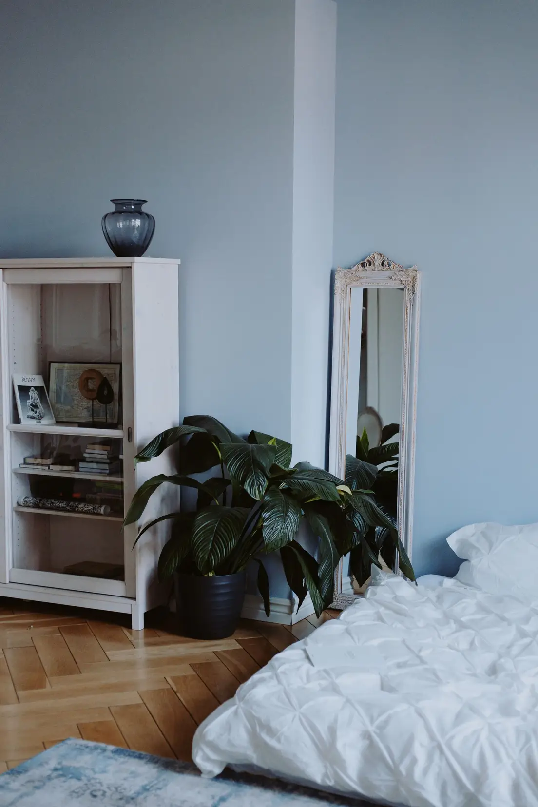A leaning floor mirror beside a potted plant and bookcase in a blue bedroom