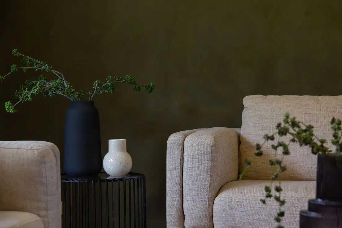 Two oatmeal textured armchairs against an olive green limewash wall with a black vase and a speckled ceramic vase