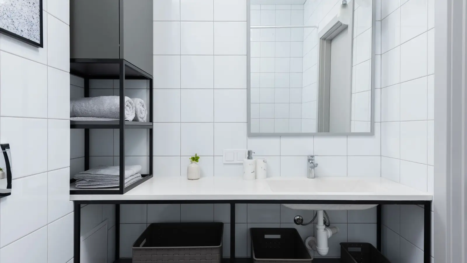 Black open wall shelving holding rolled towels above a small bathroom counter with storage baskets