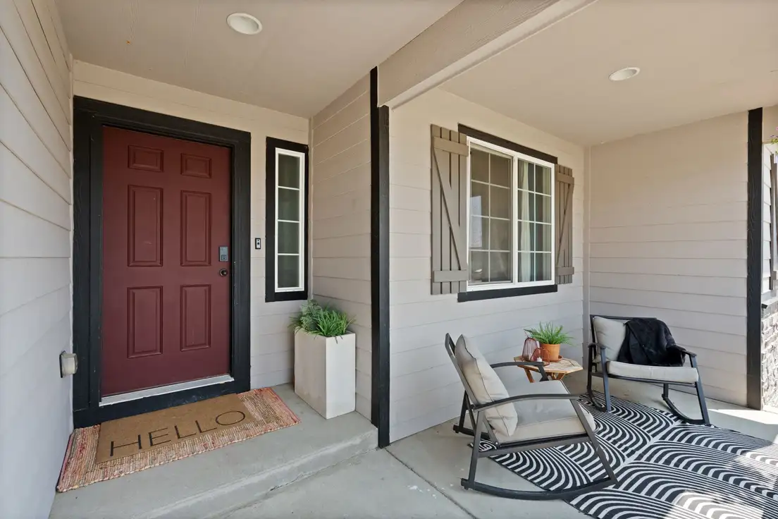 A covered small front porch with a red door, a coir doormat, and a black and white outdoor rug under two rocking chairs