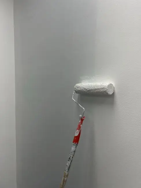 A close-up of a paint roller applying smooth white paint to a bedroom wall, showing the sheen finish on bedroom walls