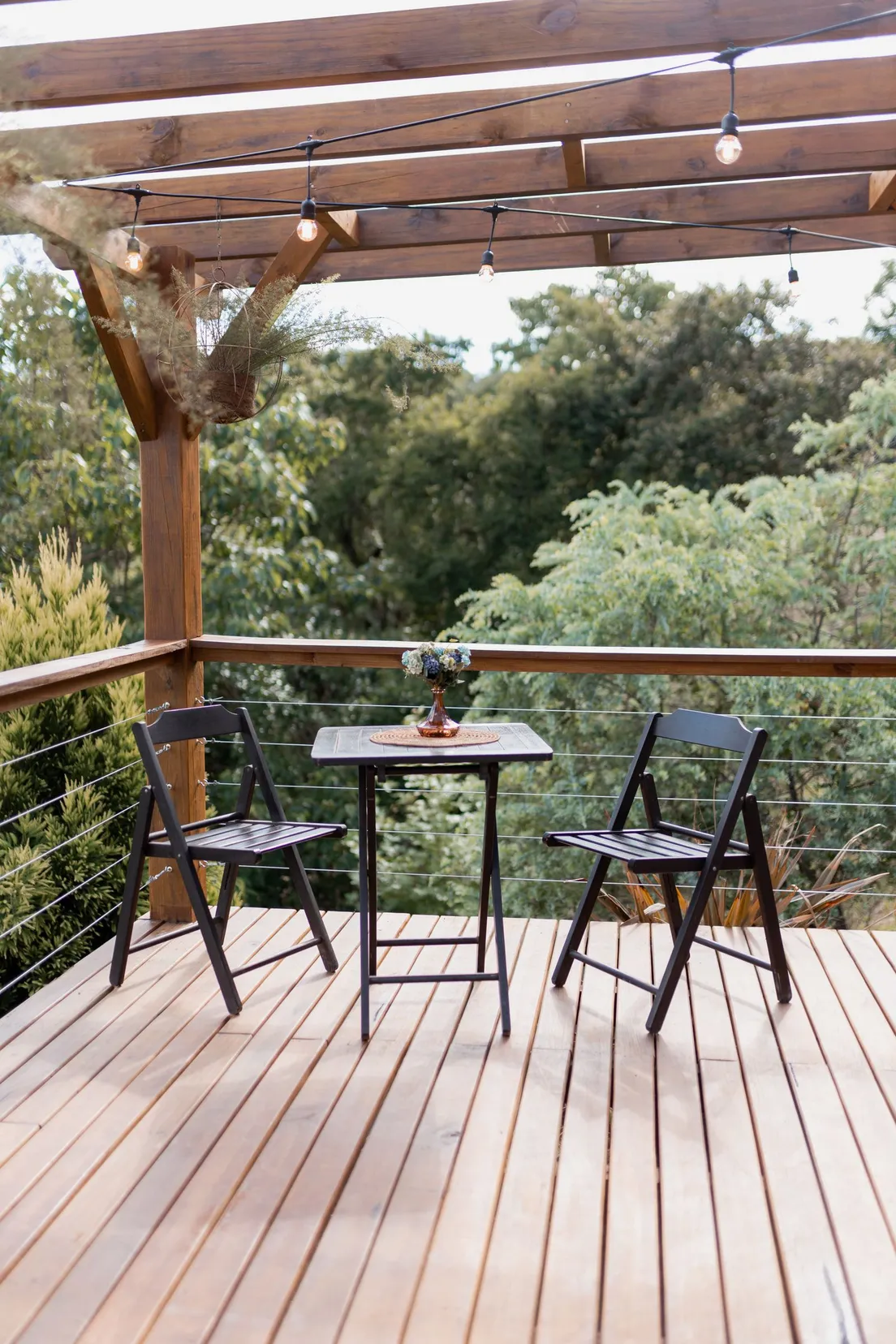 A small wooden pergola with string lights over a bistro table and two chairs on a patio