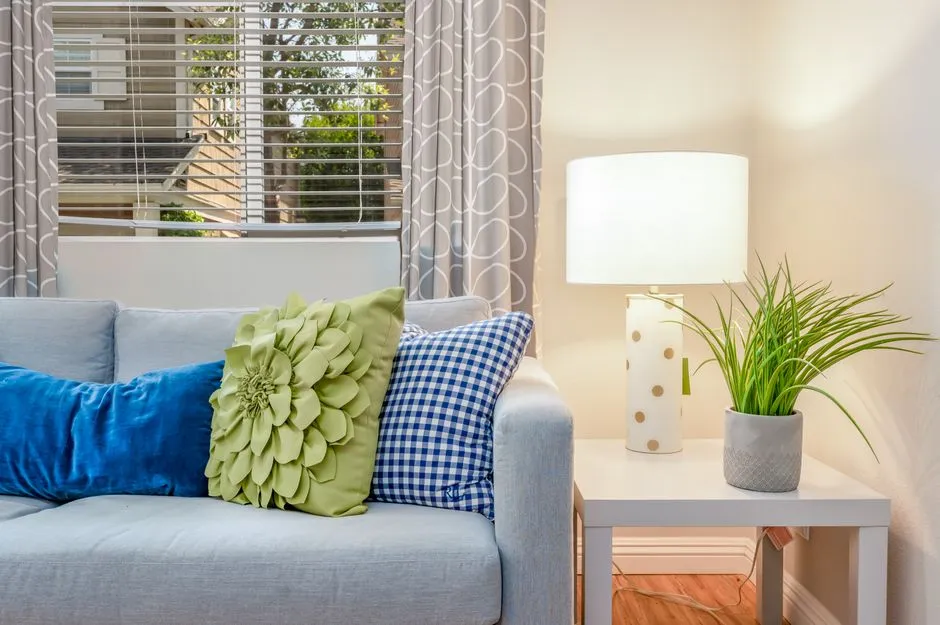A gray couch with blue, green floral, and blue gingham throw pillows beside a lamp and a potted plant