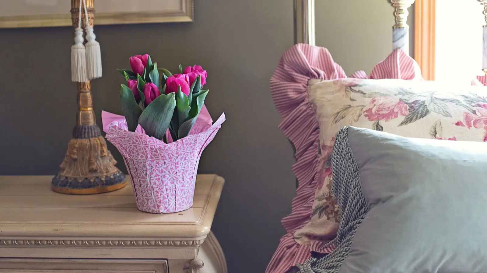 Pink tulips wrapped in patterned paper on a nightstand beside a ruffled floral bed pillow