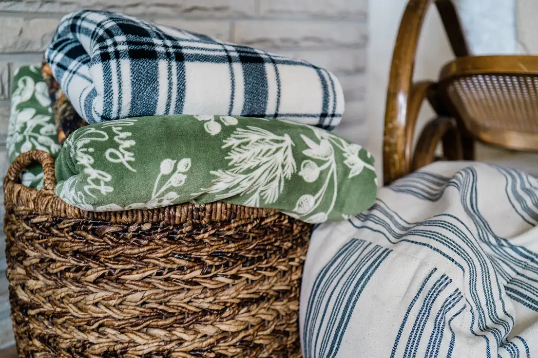 Folded plaid, botanical, and ticking-stripe throw blankets in a woven basket beside a bentwood chair, farmhouse living room textiles