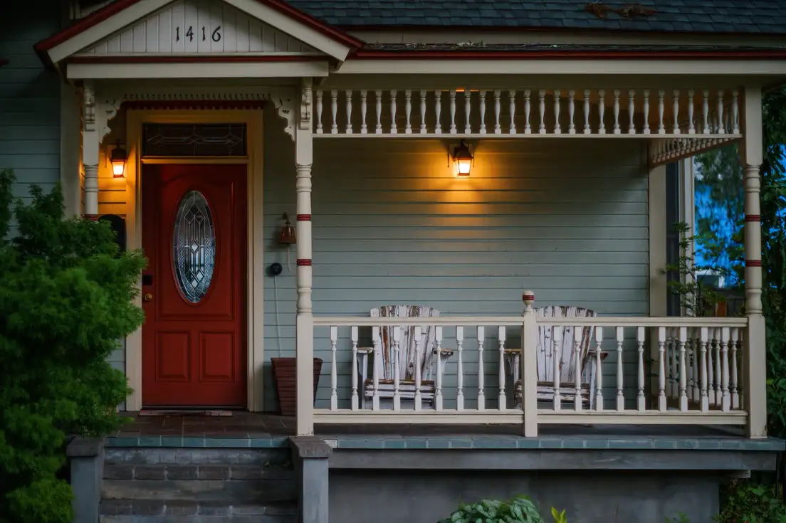 A Victorian front porch at dusk with two lit lantern light fixtures beside a red front door and wood porch chairs