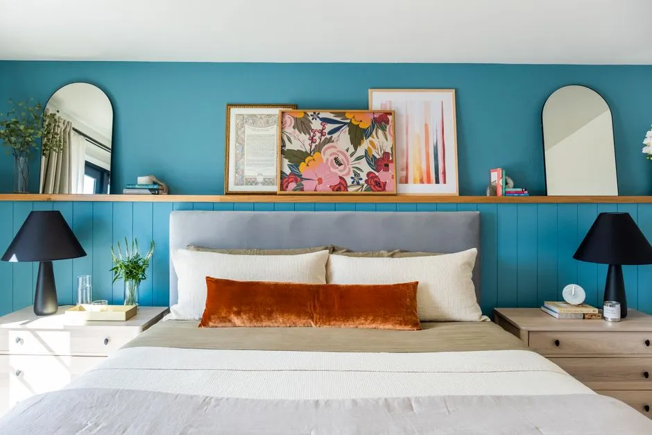 A teal bedroom wall with leaning framed art and mirrors displayed on a wall-mounted ledge