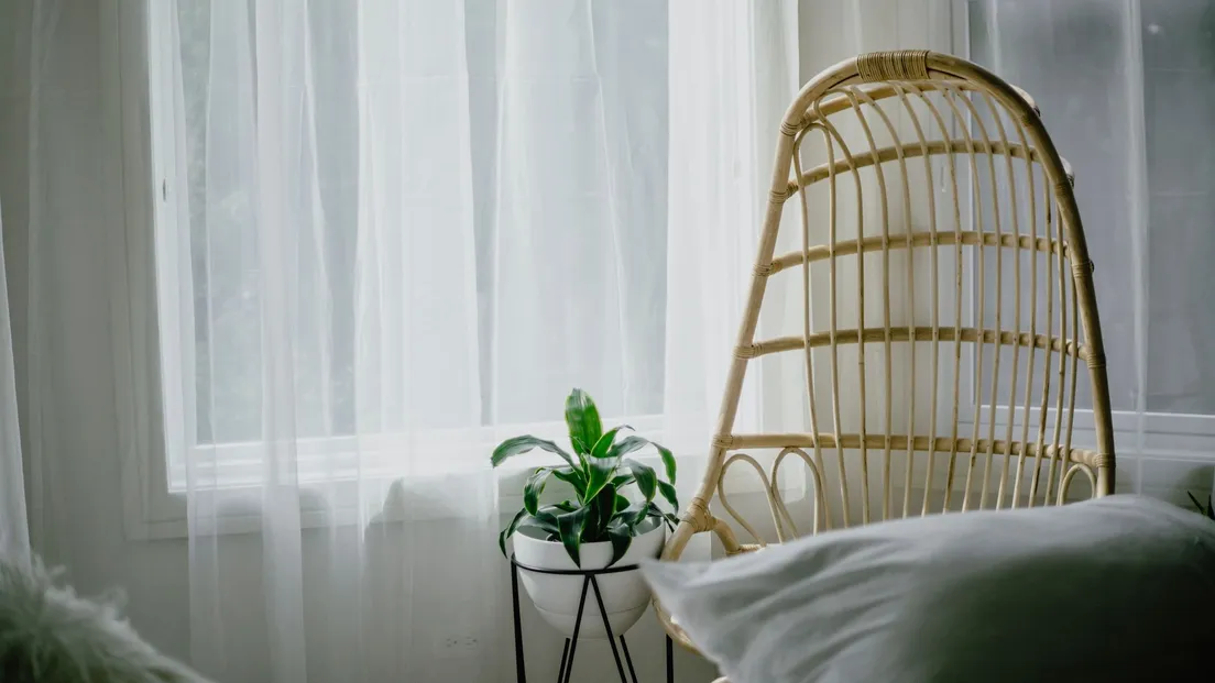 A rattan accent chair beside a sheer curtain and a potted plant in a bedroom corner