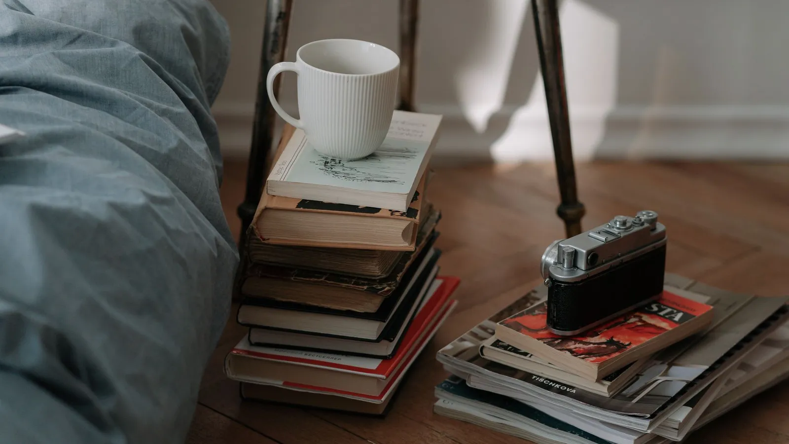 A quiet floor-level reading corner with a stack of books, a mug, and a vintage camera beside a bed