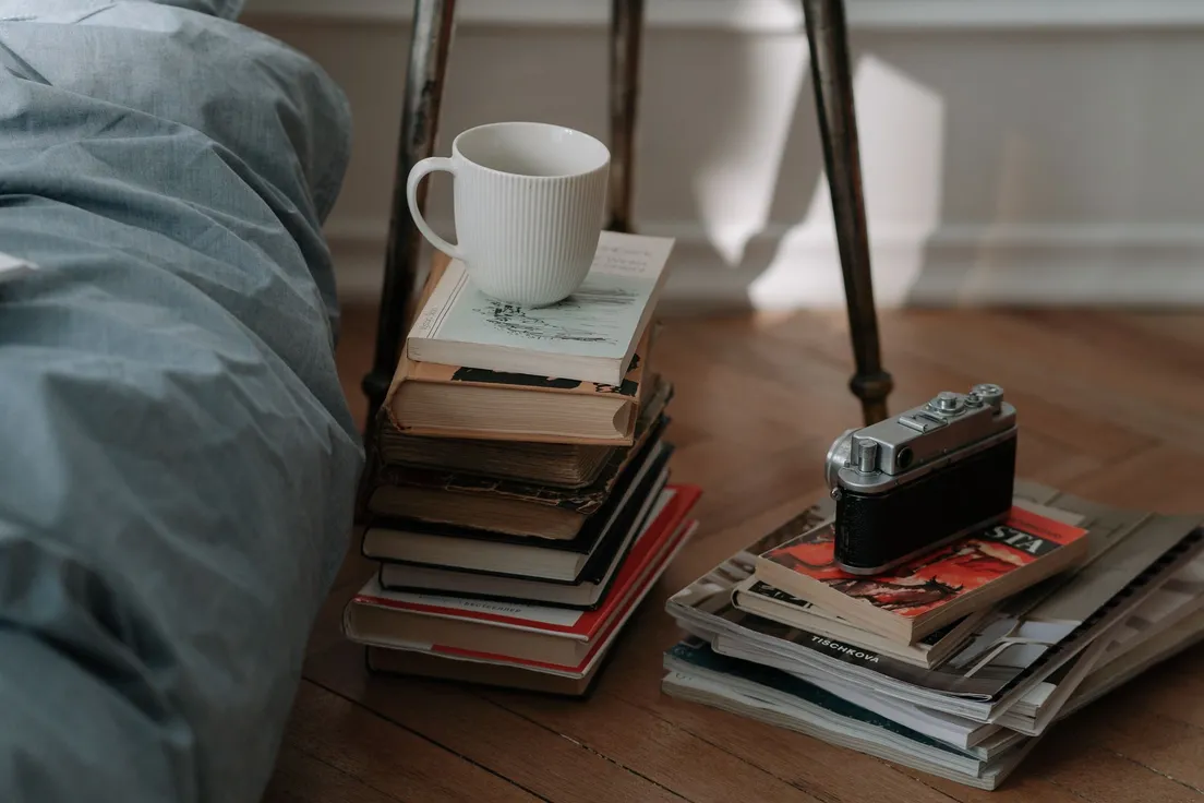 A quiet floor-level reading corner with a stack of books, a mug, and a vintage camera beside a bed