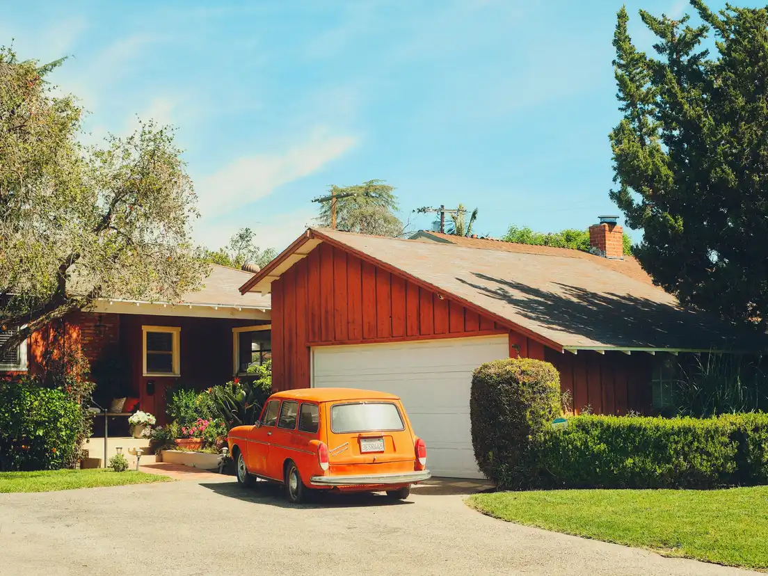 A barn-red board-and-batten ranch house exterior with a white garage door, yellow window trim, and an orange vintage car in the driveway