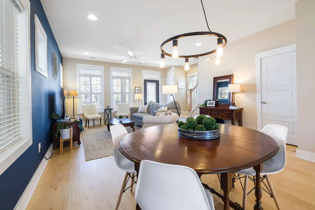 A round wood dining table with white chairs under a ring chandelier, with the living area and sofa visible beyond in a living room dining room combo