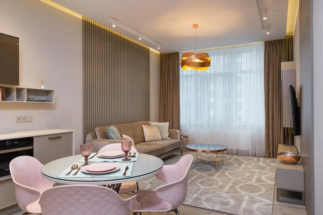 A round glass dining table with pink chairs in front of a sofa and coffee table on a patterned area rug that defines the living zone